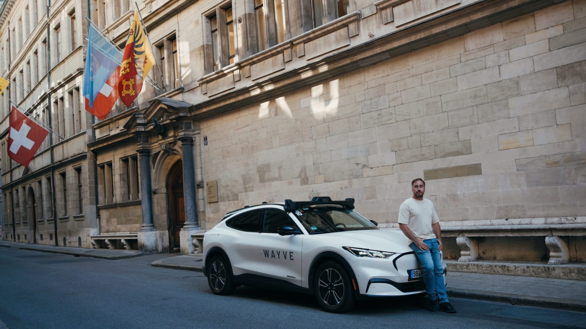 Man leaning against Wayve car with a big walled building in the background