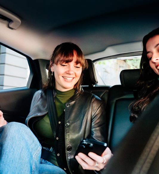 two people smiling while looking at a phone in the backseats of a car