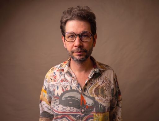 headshot of man with glasses and colourful shirt against brown background